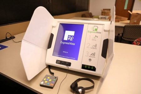 White ExpressVote voting machine with blue screen showing ES&S logo sits on white table. A game controller style tactile interface and black headphones are attached to the machine