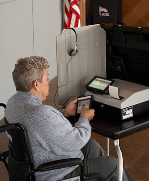 White voter with short hair sits in wheelchair in front of voting machine. Voter holds tactile controller. Headphones are attached to gray privacy screen. Behind privacy screen is American flag.