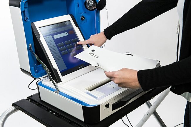 White ballot scanner with blue touch screen and blue case rests atop black platform. White hand of person wearing black shirt touches screen. Screen has number buttons and reads "Ready for voting."