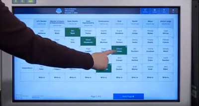 Voter with white hand and brown shirt points to white ExpressVote XL voting machine screen. Voter makes a selection that turns green