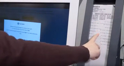 Voter with white hand and brown shirt pointing to white ExpressVote XL ballot card that appears behind plexiglass window