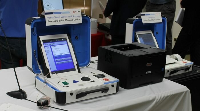 n open white and blue case is positioned on a table with a white tablecloth. Inside the case is a large blue touch screen encased in a white housing on a white base, which houses a tethered tactile interface and a small thermal printer. To the right of the unit is a square, black printer. Another unit is on a table to the right of the printer.