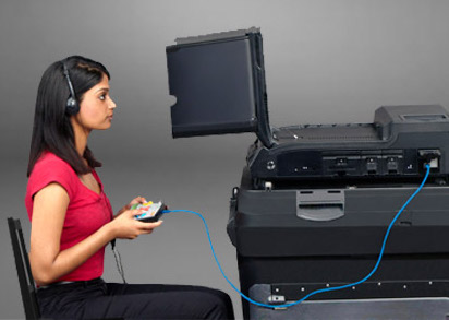 Voter with brown skin and black hair holding tactile controller tethered to ballot scanner/ballot marking device. Voter is wearing headphones and looking at a screen behind a privacy partition