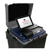 Black scanner with white touch screen reading "FreedomVote. Unisyn." Scanner has gray ballot slot and small black receipt printer below screen. Base of scanner is black.