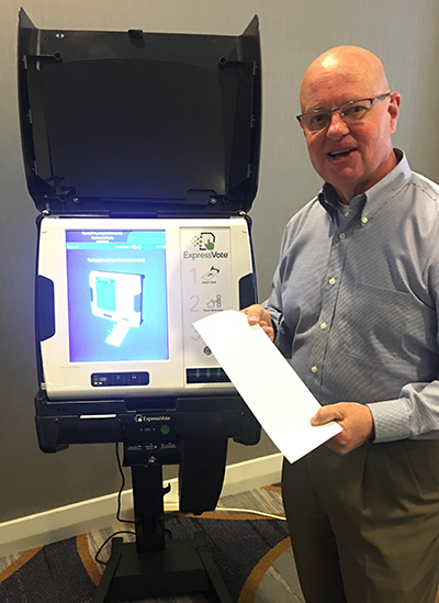White man with bald head and glasses wears a blue collard shirt and holds a blank white ballot card next to an ExpressVote voting machine with a blue and white screen that displays an image of the machine. The voting machine is white and sitting atop a black rolling cart with a black hood