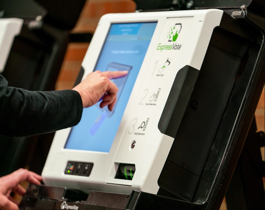 Voter with white hand and black sweater touches screen of a white ExpressVote voting machine