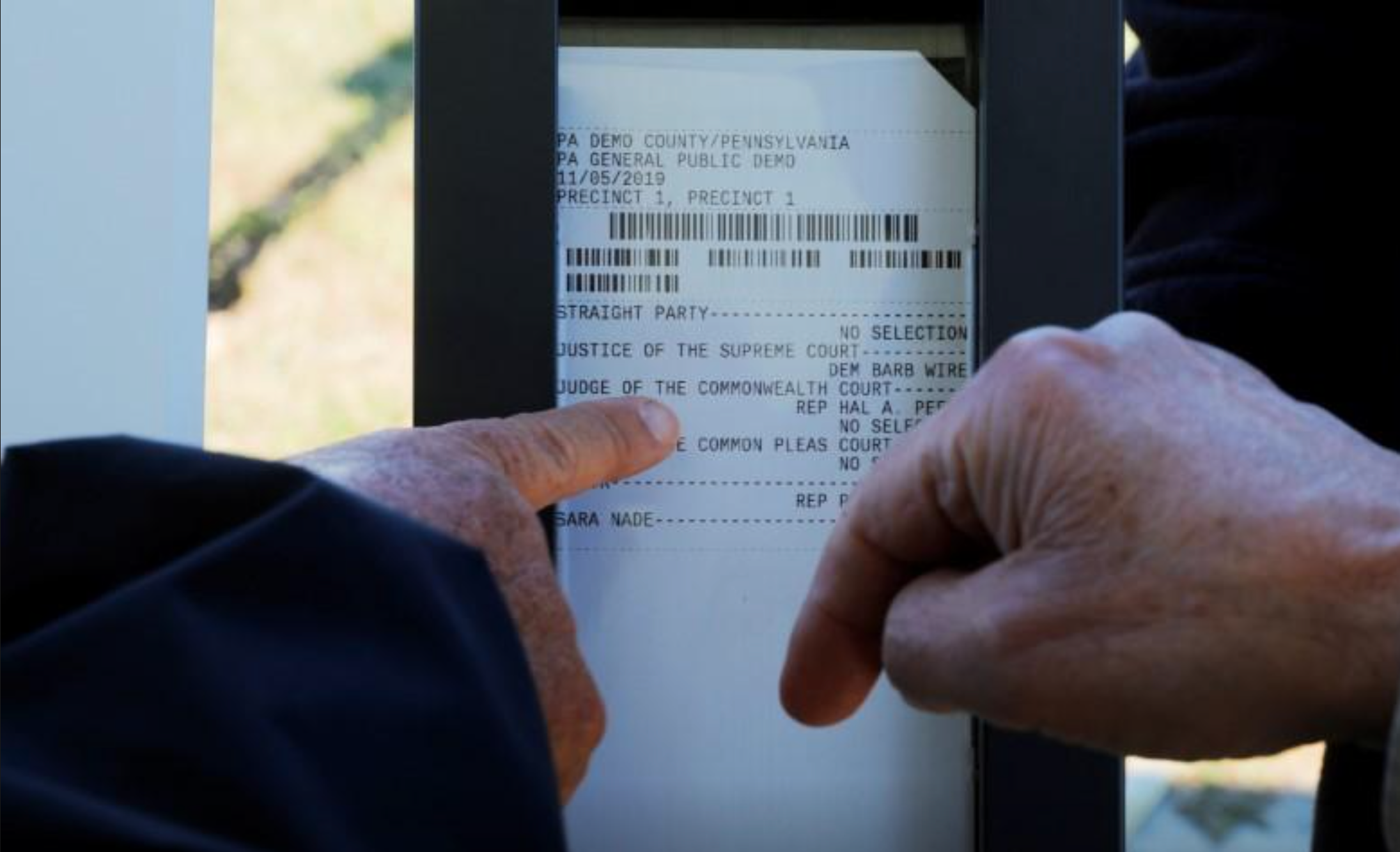 Two white hands point at a ballot card behind a plexiglass window