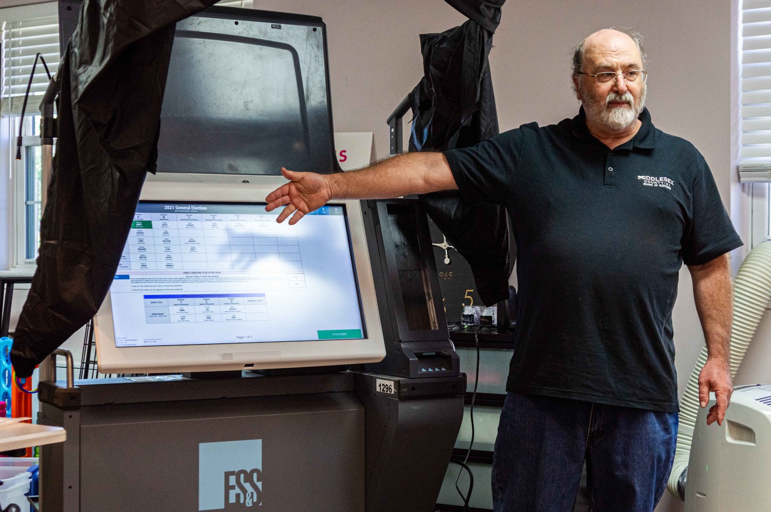 While man with glasses and white beard points to ExpressVote XL voting machine on a black cart