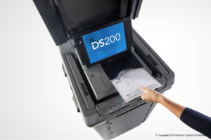 Gray scanner with blue screen reading "DS200." White hand inserts ballot into scanner.