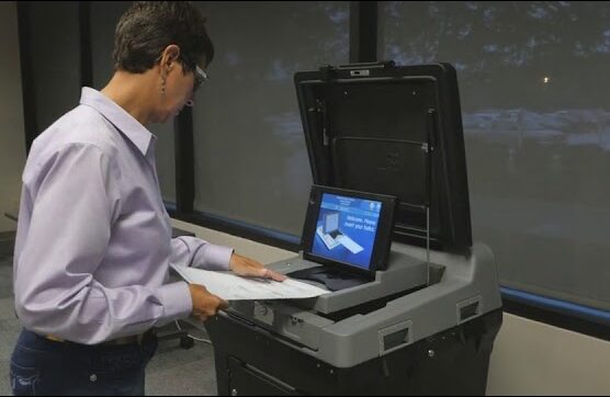 Election worker wearing lavender shirt and jeans demonstrates how to insert ballot into gray scanner on black base. Scanner has blue screen that reads "Welcome, please insert your ballot."