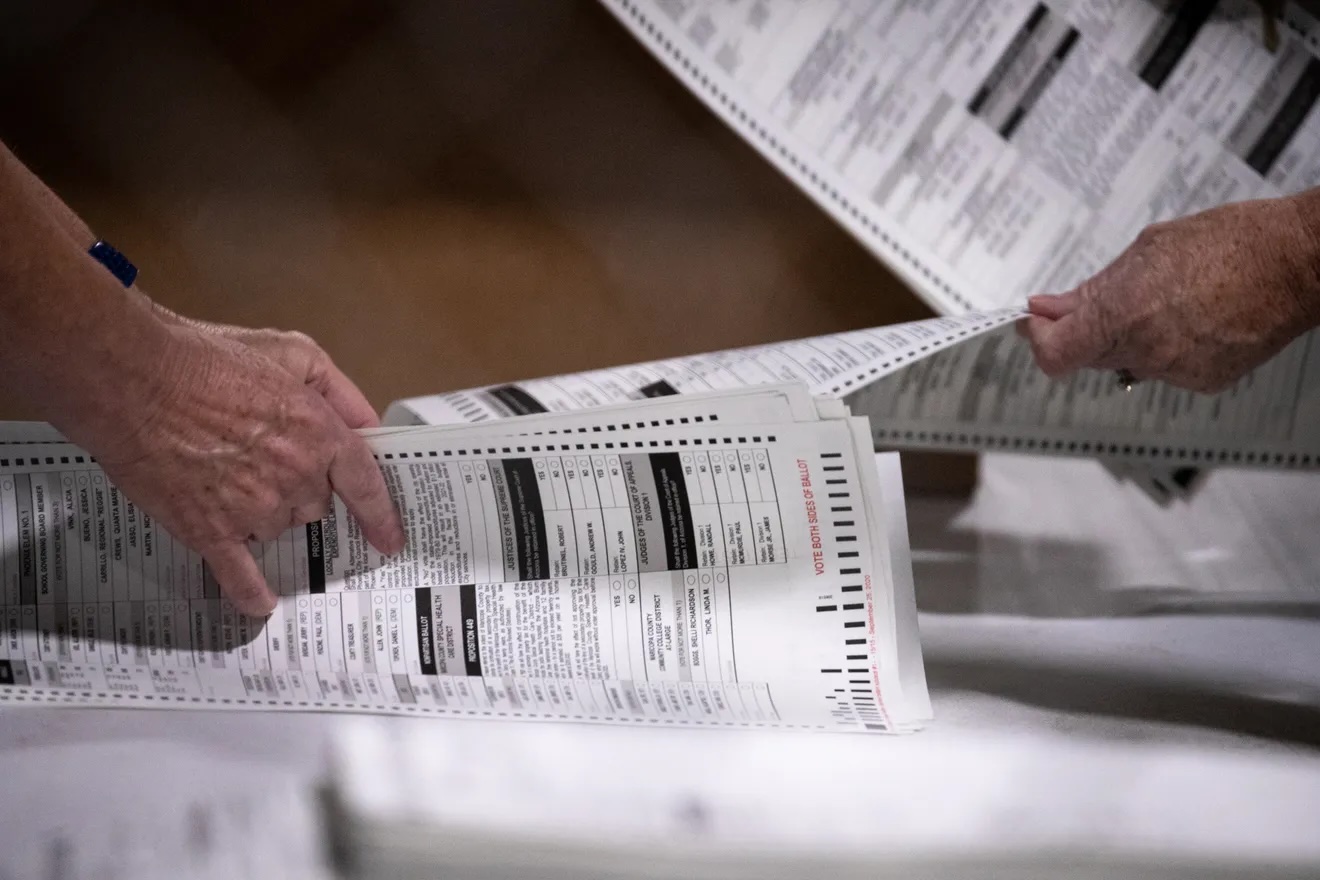 The hands of two white humans are shown sorting through paper ballots.