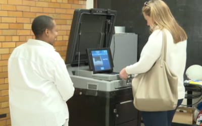 White voter with blond hair scans ballot into black and gray ballot scanner with blue screen. Black poll worker stands nearby smiling