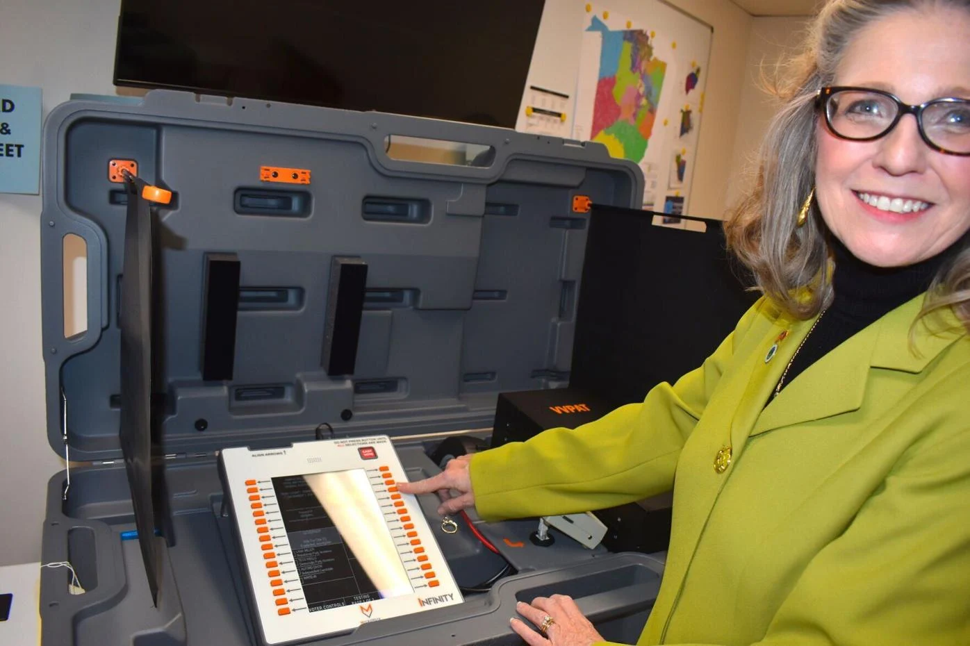 White woman wearing glasses and green blazer shows push buttons on white voting machine in gray case