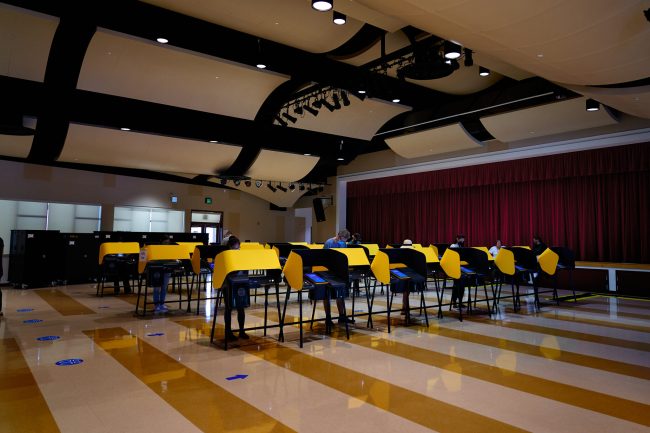 There is a lineup of yellow polling booths inside an auditorium. The screens are blue and voters stand at some of the booths.
