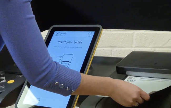 VSAP_insert ballot Voter wearing blue shirt inserts their blank ballot into a scanner. To its left is a touchscreen that reads "insert your ballot"