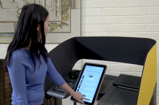 VSAP_review ballot Voter with black hair and blue shirt touches a screen that reads "review your selections." She is standing in front of a yellow polling booth.